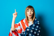 © Alex - Young woman wrapped in USA flag with a surprised face on a blue background. USA visa concept, English language, gesture is all right, thumbs up, like