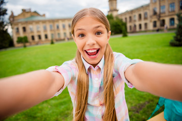 Wall Mural - Close up photo of cheerful kid with pigtails screaming laughing making photo wearing checkered t-shirt outside academia