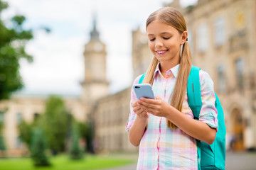 Wall Mural - Portrait of concentrated small girl using device holding rucksack backpack wearing checkered plaid t-shirt standing outside