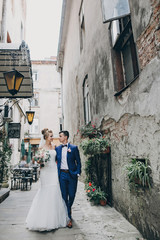 Wall Mural - Stylish happy bride and groom walking in old city street. Gorgeous wedding couple of newlyweds embracing and smiling outdoors. Romantic moment