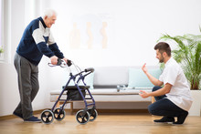 Elderly Grandfather With Walker Trying To Walk Again And Helpful Male Nurse Supporting Him
