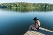 © Hernandez And Sorokina/ADDICTIVE STOCK - Side view of young woman in black swimsuit and hat sitting on wooden pier on the mobile phone on a lake on clear blue sky and forest background