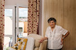 © Patricia Becaroto/ADDICTIVE STOCK - Elderly curly gray haired female in white shirt with hand on waist standing while leaning on armchair at home and looking at camera
