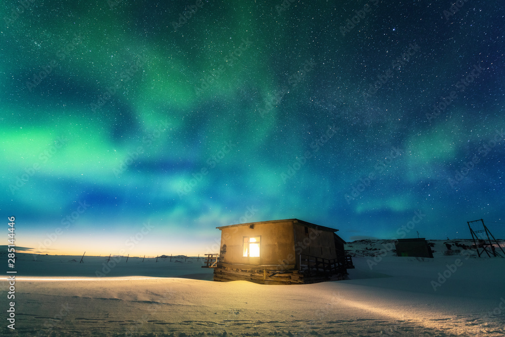 Aurora borealis over old small house with yellow light from the window ...