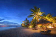 Sandy Beach With Palm Trees And Sunbeds Under Umbrellas During Blue Hour In Summer.  Landscape With Sea Shore, Beautiful Starry Sky, Green Palms And Yellow Light At Night. Travel In Zanzibar, Africa