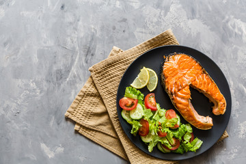 Fried salmon fish steak with vegetable salad and lemon on black plate. Creative layout made of fish, lettuce, tomato, cucumber, top view, flat lay, mockup, overhead. Healthy food concept