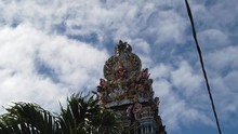 A Shot Of The Tower Of A Hindu Temple Full Of Deities As Walking Down The Street, With A Palm In The Middleground And Many Tiny Clouds In The Sky