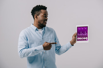 happy african american man pointing with finger at digital tablet with medical app on screen isolated on grey