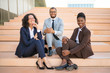© Mangostar - Happy business team posing for camera near office building outside. Cheerful multiethnic man and women sitting on stairs and smiling. Successful team portrait concept