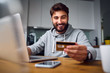 © baranq - Portrait of smiling young man making online payments from home with credit card and laptop