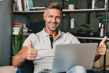 Sticker - Photo of unshaven adult man smiling and showing thumbs up while using laptop in apartment