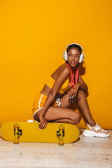 Wall Mural - Image of adorable african american woman listening to music with headphones while sitting on skateboard