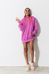 Poster - Positive optimistic happy young african stylish woman in pink sweatshirt and sunglasses posing isolated over white wall background showing peace gesture.