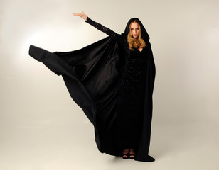 Wall Mural - full length portrait of blonde woman wearing long black flowing clock and lace gown, standing pose against a creamy studio background.