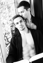 Male Gay couple sitting in window reading to each other, one holds book with partner kissing his hair