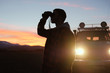 © Sibstock/Stocksy - A man with binoculars near the car at sunset. Man Binoculars Looking Mountain Cloudscape Traveling Concept