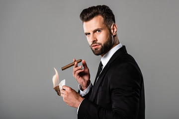 side view of rich businessman in black suit lighting up cigar from burning dollar banknote isolated on grey