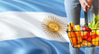 Woman is holding supermarket basket, Argentina waving flag background. Economy concept for fresh fruits and vegetables.