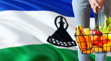 Woman Is Holding Supermarket Basket, Lesotho Waving Flag Background. Economy Concept For Fresh Fruits And Vegetables.