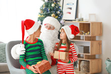 Santa Claus And His Little Helpers With Gifts In Room Decorated For Christmas