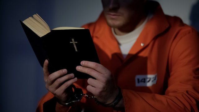 Imprisoned man in handcuffs reading bible, feeling guilty and praying for soul