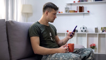 Relaxed Male Soldier Typing On Smartphone, Sitting On Sofa With Tea Cup At Home
