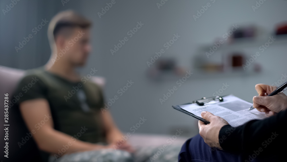 Psychologist making notes during therapy session with sad male soldier, PTSD - obrazy, fototapety, plakaty 