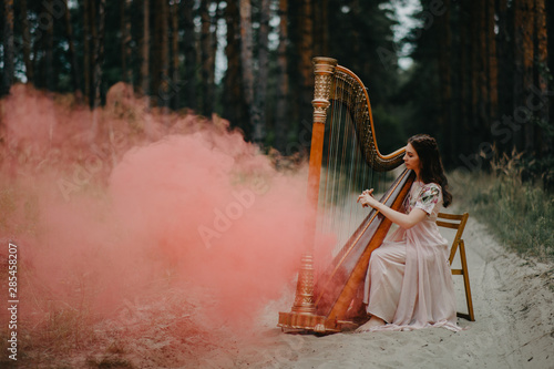 Fotografía Woman harpist sits at forest and plays harp against a background of pines and smoke
