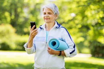 fitness, sport and healthy lifestyle concept - happy smiling senior woman with exercise mat and smartphone at park