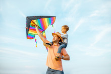 Portrait Of A Happy Father And Young Son On The Shoulders With Colorful Air Kite On The Blue Sky Background. Concept Of A Happy Family And Summer Activity