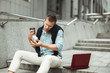 © studioprodakshn - young handsome office worker having business conversation while eating his sandwich and drinking coffee for lunch during break sitting on the stairs outside with laptop
