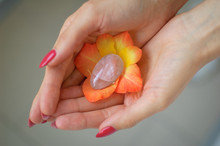 Female Hand With Red Manicure Holding Transparent Violet Amethyst Yoni Egg For Vumfit, Imbuilding Or Meditation Inside Orange And Yellow Gladiolus Flower.