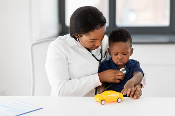 Poster - medicine, healtcare, pediatry and people concept - african american female doctor or pediatrician with stethoscope listening to baby patient on medical exam at clinic