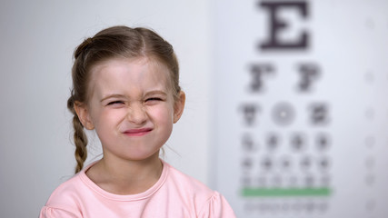 Wall Mural - Cute girl screwing up her eyes, trying to see letters on table for vision test