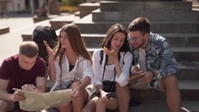 Four Young Friends Are Sitting On A Stairs Of Some Ancient Building And Looking At The Map Of The City. They Are Tourists And They Are Deciding The Way Where To Go.