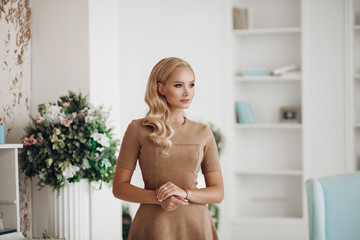 Gorgeous pretty lady sitting in luxury apartment and holding hands on knees. Beautiful young woman in elegant beige dress posing on white sofa. Happy blonde model relaxing after long working day.