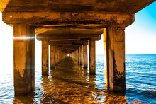 Diminishing Perspective Of Wooden Pillars Underneath Long Old Pier Standing In Ocean Water At Sunset