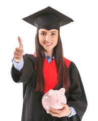 Wall Mural - Female graduate with piggy bank showing thumb-up on white background. Tuition fees concept