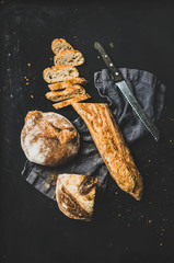 Flat-lay of freshly baked baguette cut into slices and loaf on dark linen over rustic black background, top view