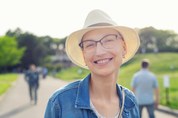 Happy young caucasian bald woman in hat and casual clothes enjoying life after surviving breast cancer. Portrait of beautiful hairless girl smiling during walk at city park after curing disease