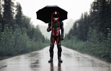Motorcyclist In Full Gear And Helmet With Umbrella In The Rain. Motorcyclist In The Dark Woods