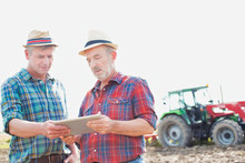 Senior Farmer Using Digital Tablet While Talking To Mature Farmer Standing Against Tractor In Field