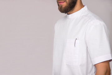 close-up of young stylish bearded nurse man in white medical shirt with pen in pocket which is standing and smiling on white wall background. medical fashion concept. free space on left side