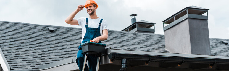 Wall Mural - panoramic shot of happy repairman sitting on roof and holding toolbox