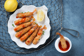 Plate of whole deep-fried shrimps in breadcrumbs, flatlay over blue stone background with a fishing net, horizontal shot