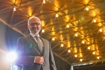 Wall Mural - Low angle view of senior businessman in suit wearing eyeglasses standing against illuminated roof