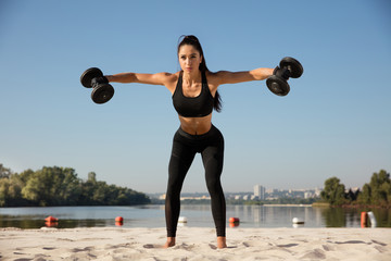 Young healthy woman training upper body with weights at the beach. Single caucasian female model practicing at the river side in sunny day. Concept of healthy lifestyle, sport, fitness, bodybuilding.