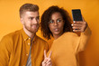 © antianti - Studio shot of overjoyed guy laughs sincerely while poses for selfie with girlfriend, she shows victory or peace sign, have fun together, use modern cell phone for entertaining, isolated yellow wall