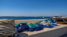 View On The Fishing Gear And Equipment On The Port On Blue Sky Background. Lay Out Various Fishing Nets, Hawser, Mooring Line, Ropes. Preparing To Sail. Drying And Repairing Nets On The Dock.