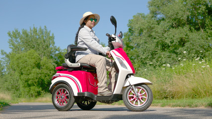 Portrait of an African American woman in sun glasses and hat riding electric scooter. Freedom of riding mobility vehicle along green city park on summers day.